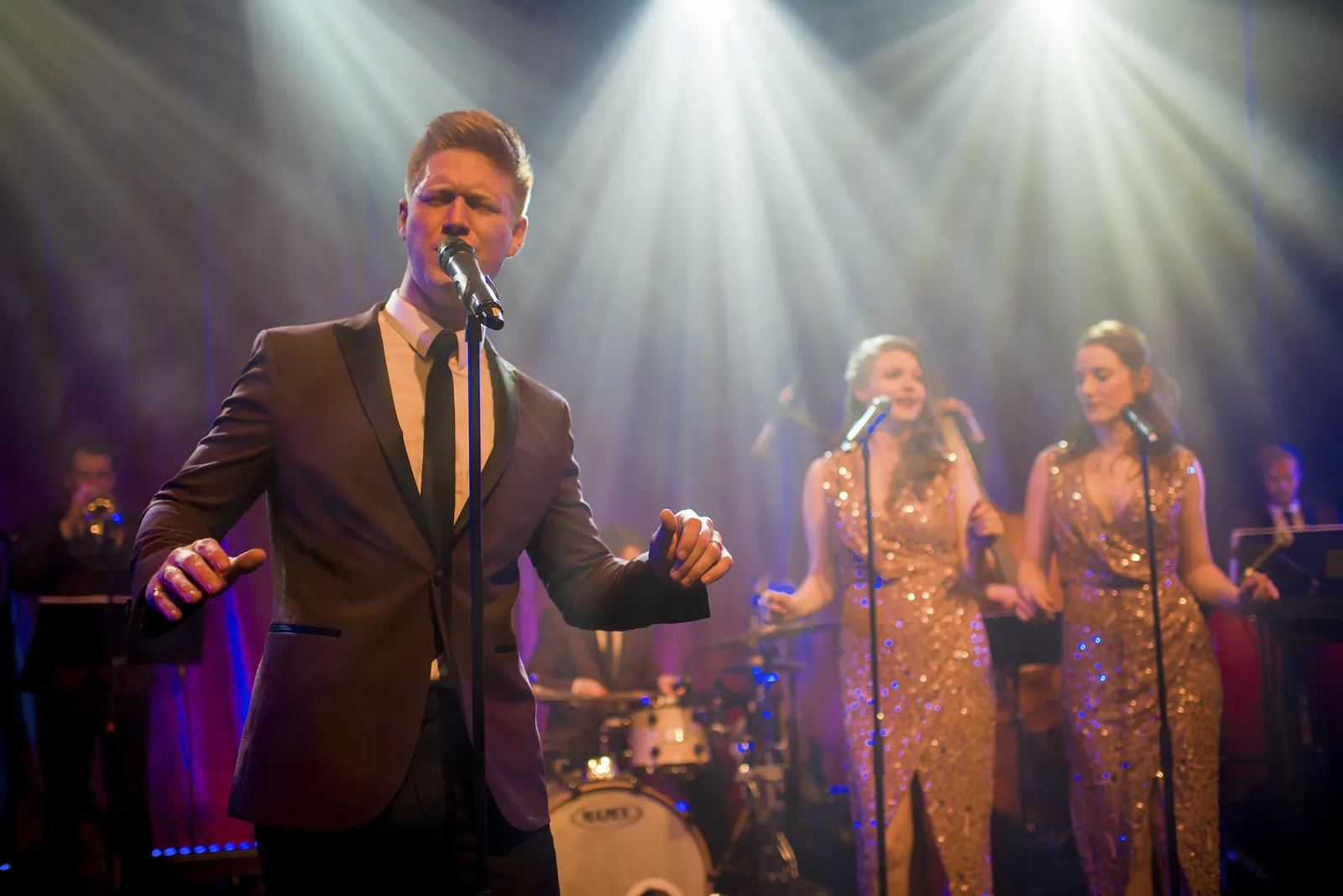 A male singer in a suit performs on stage under bright lights with a jazz band, whilst two female backing singers in sparkling dresses and musicians play instruments in the background.