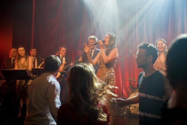 A jazz band performs energetically on stage with a female lead singer under dramatic stage lights, whilst people in the foreground dance and enjoy the lively atmosphere.