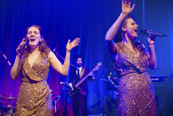 Two women in gold sequin dresses sing passionately on stage as part of a jazz band, holding microphones, with a guitarist in a suit playing in the background under blue and purple stage lights.