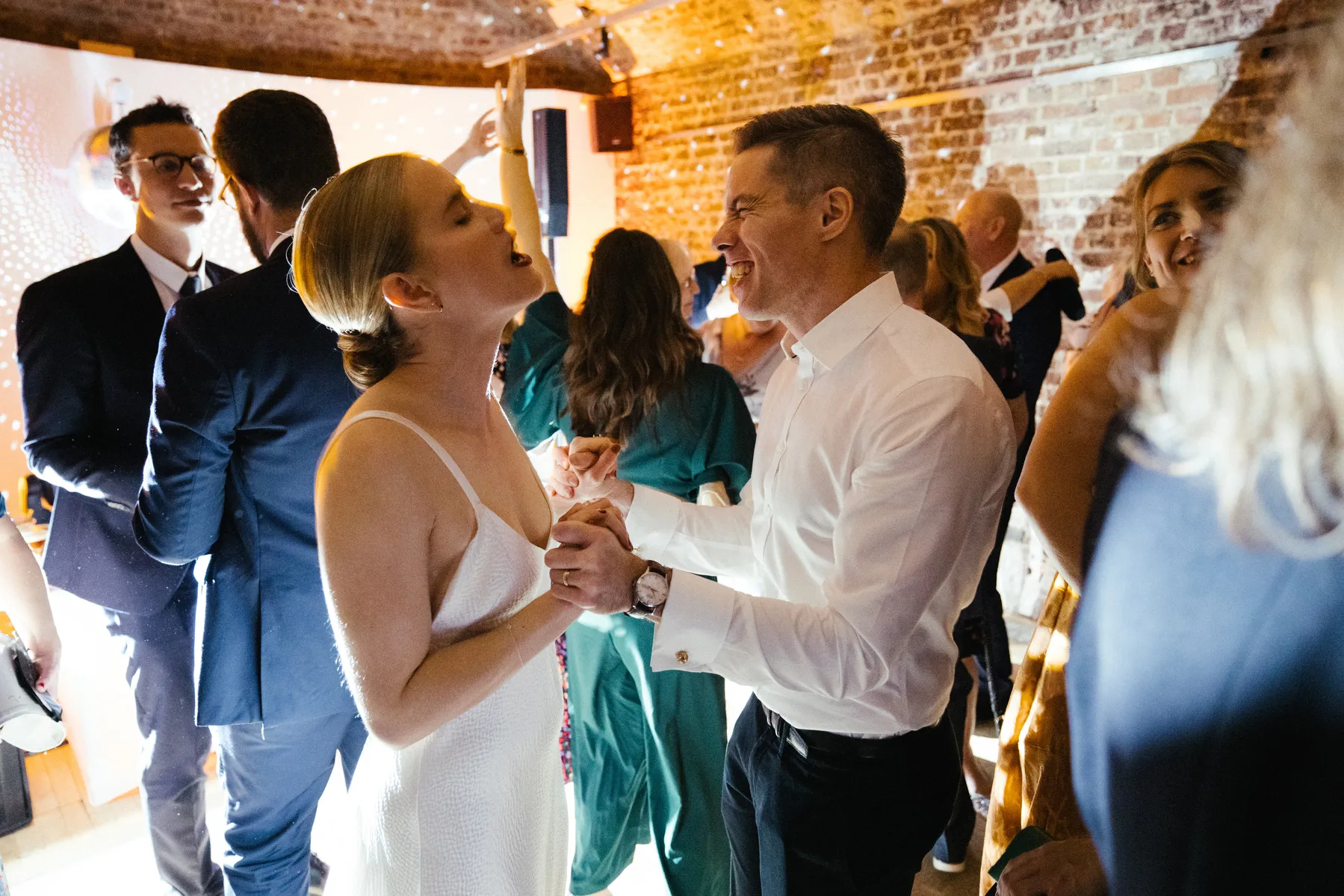 A bride in a white dress and a man in a white shirt hold hands and smile at each other whilst dancing at a lively indoor RSA house wedding reception with other guests in the background.