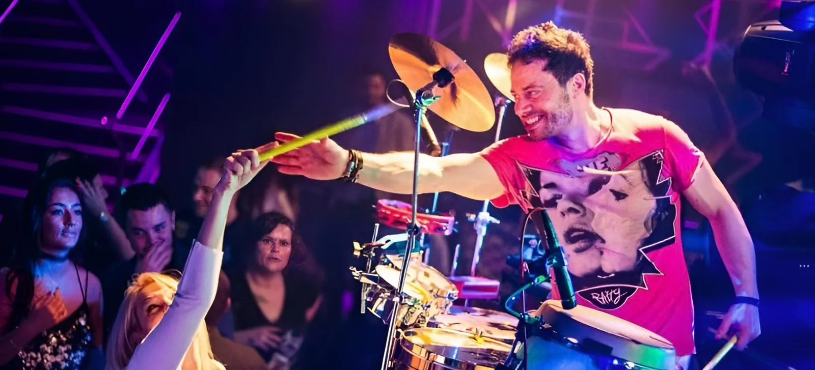 A smiling percussion drummer on stage, wearing a red graphic t-shirt, reaches out with a drumstick towards an enthusiastic audience member. The crowd looks engaged and colourful lights illuminate the scene.
