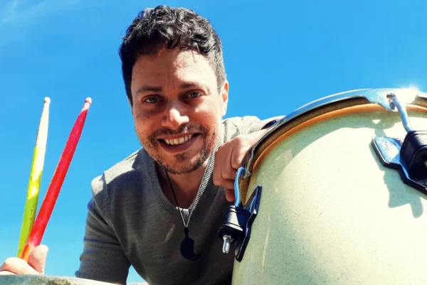 A smiling man holds two colourful percussion drumsticks next to a large drum under a clear blue sky, looking directly at the camera.
