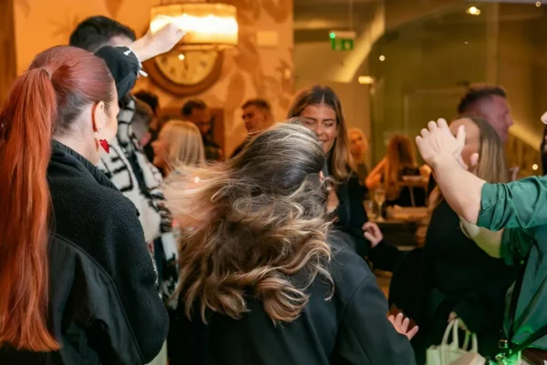 A group of people are smiling, laughing, and dancing together indoors at a lively social event, with upbeat percussion music adding energy to the warm lighting and festive decor for a cheerful, celebratory atmosphere.
