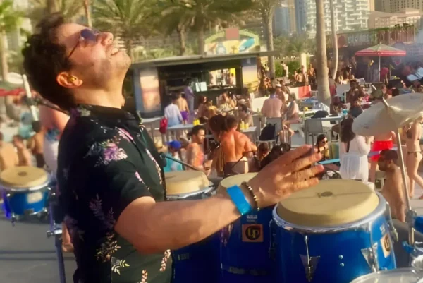 A musician wearing sunglasses and a floral shirt plays blue percussion conga drums on an outdoor stage. Behind him, a lively crowd enjoys a sunny day at a beachside venue lined with palm trees.
