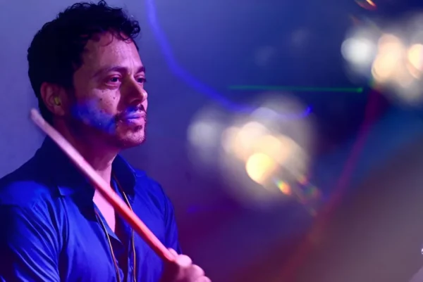 A man wearing a blue shirt plays percussion drums under colourful, moody stage lighting, with reflections and bokeh effects in the foreground.