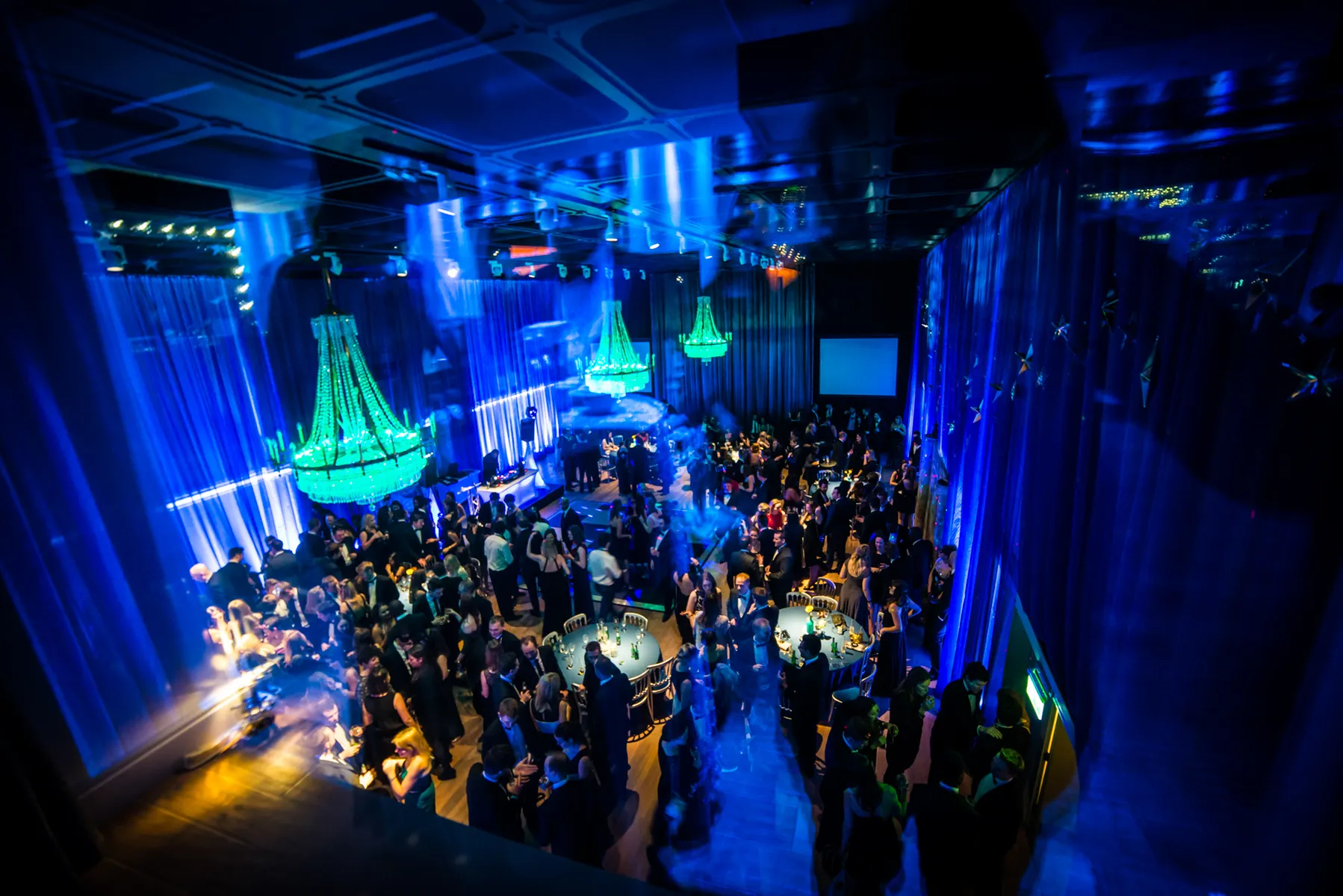 A large crowd in formal attire gathers in a dimly lit ballroom with blue lighting, green chandeliers, and draped curtains, as a Christmas party DJ sets the mood for an elegant, festive atmosphere.