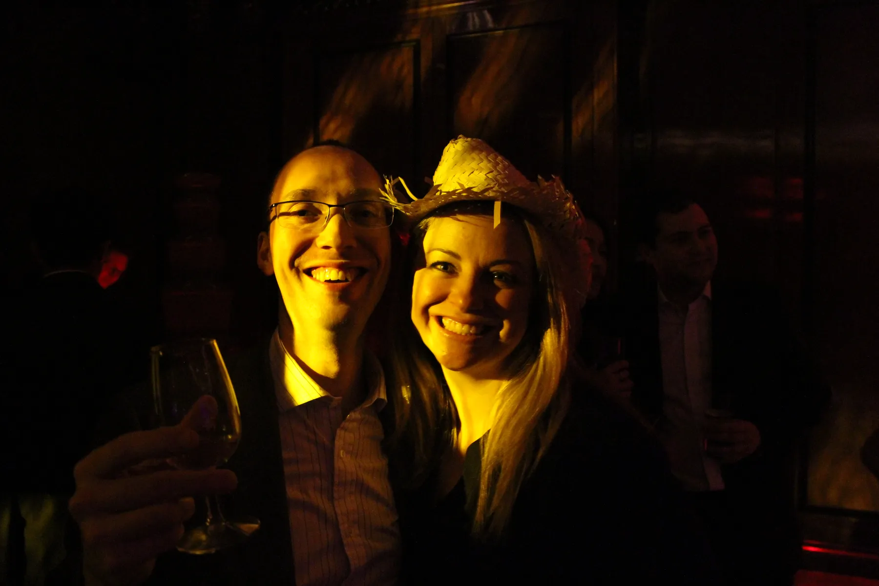 A smiling man holding a drink stands next to a smiling woman in a straw hat at a dimly lit Christmas party, with other guests and dark wooden walls in the background. Warm yellow light and music from a DJ highlight their cheerful faces.