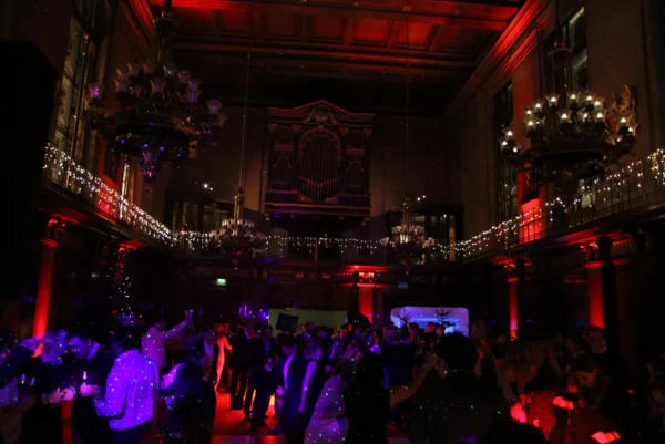 A large group of people dance in a grand, dimly lit hall with high ceilings, chandeliers, and decorative lights as a Christmas party DJ sets the mood with red and purple lighting, creating a vibrant festive atmosphere.