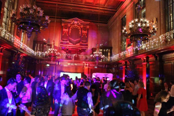 A large, ornate ballroom filled with people dancing under colourful lights. Elegant chandeliers hang from the high ceiling, and a grand organ is visible at the back. A Christmas party DJ sets the festive mood with lively music and sparkling decorations.