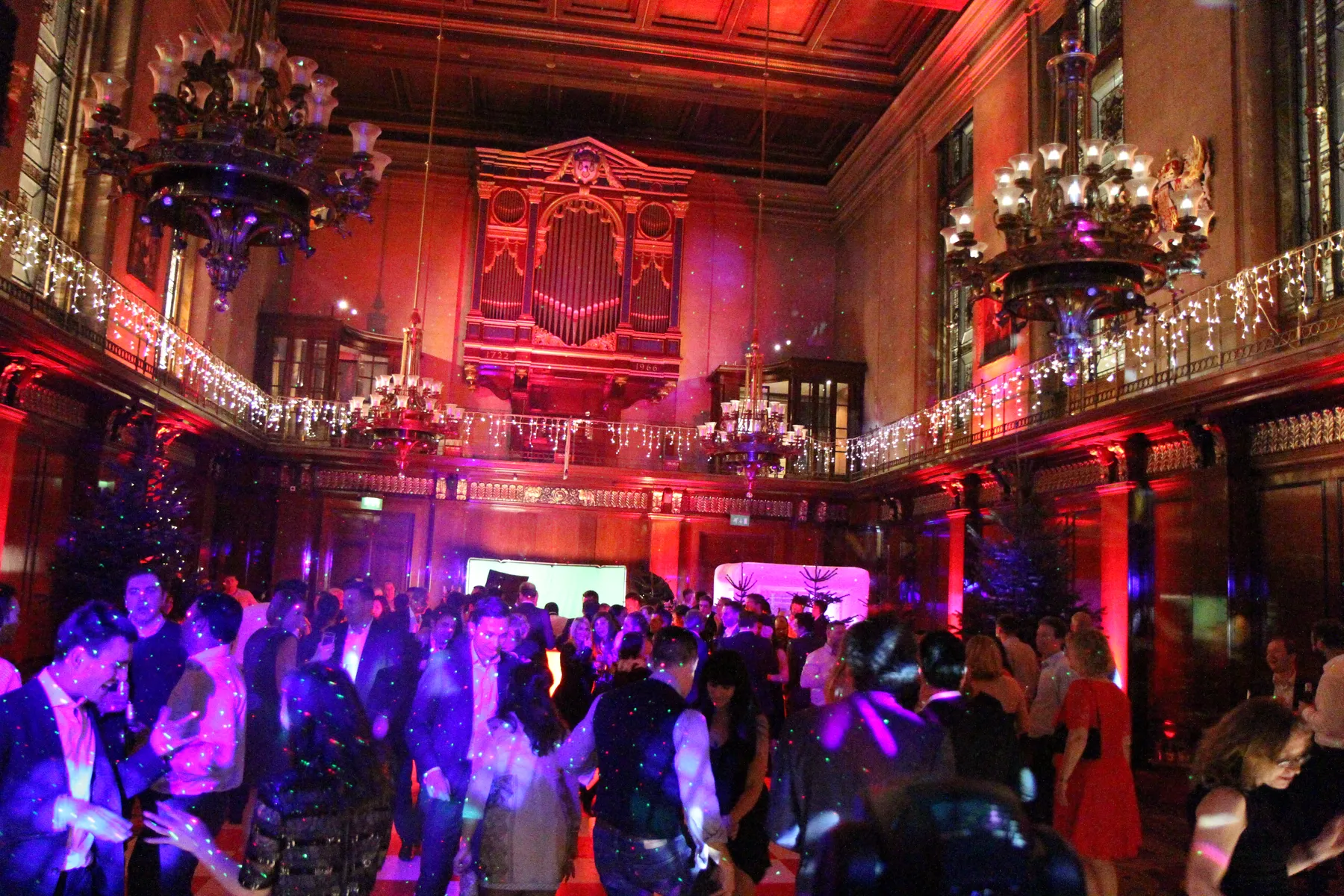 A large, ornate ballroom filled with people dancing under colourful lights. Elegant chandeliers hang from the high ceiling, and a grand organ is visible at the back. A Christmas party DJ sets the festive mood with lively music and sparkling decorations.
