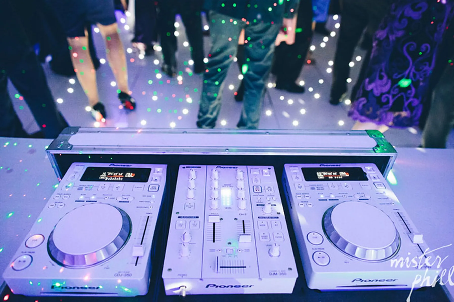 A DJ setup with mixers and turntables on a table, surrounded by colourful lights and dancing people at a lively Christmas party DJ event.