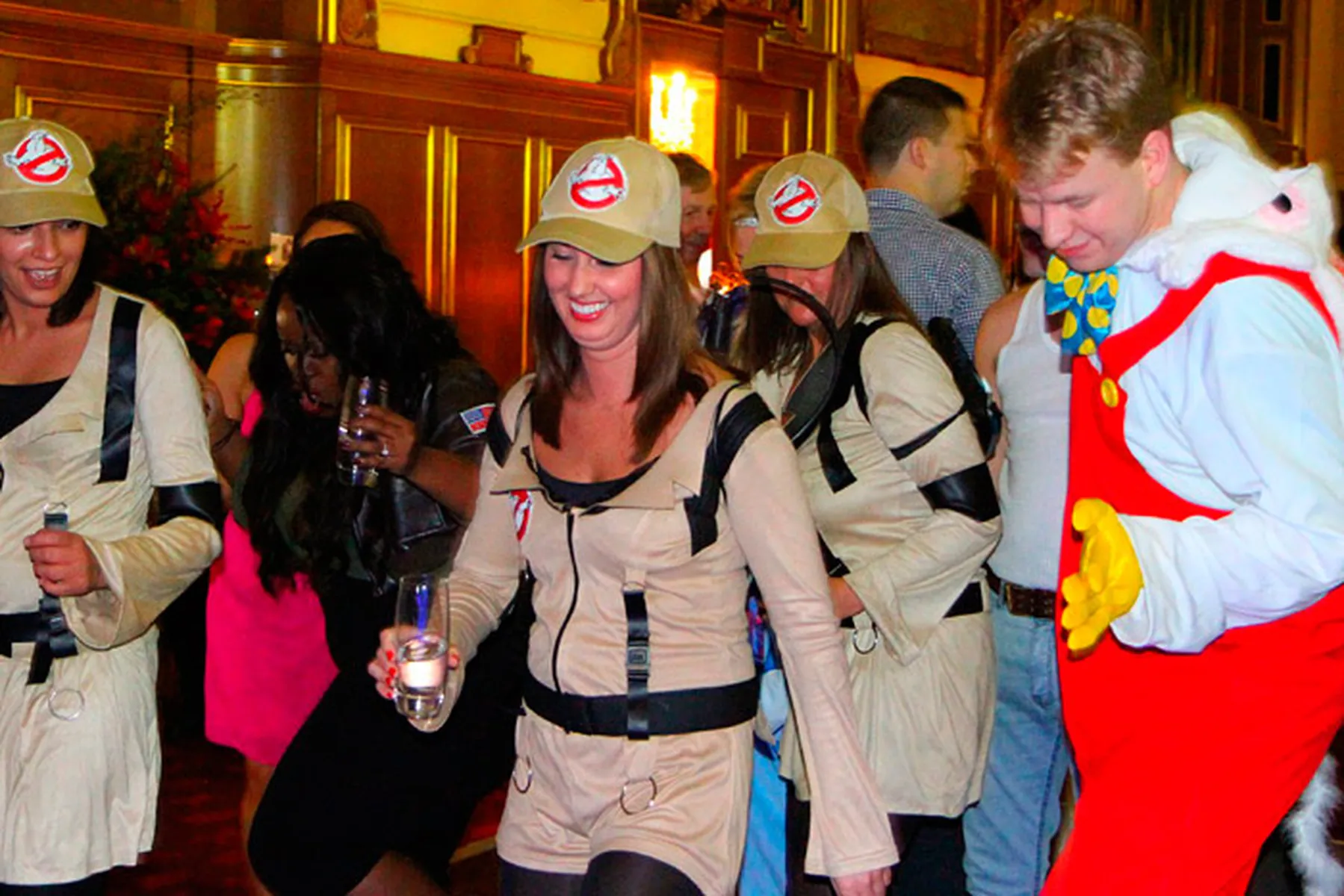 A group of people at a lively Christmas party, with three dressed as Ghostbusters and one as a cartoonish red and white animal character, all smiling and dancing together indoors to the music of a festive DJ.