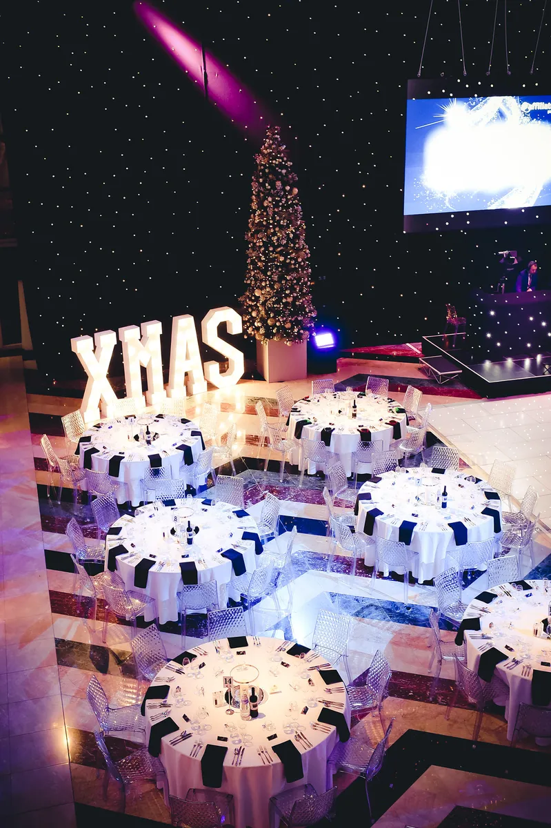 A festive banqueting hall with round tables set for dinner, a tall decorated Christmas tree, large illuminated letters spelling XMAS, a starry backdrop with a large screen, and space for a Christmas party DJ to set the mood.