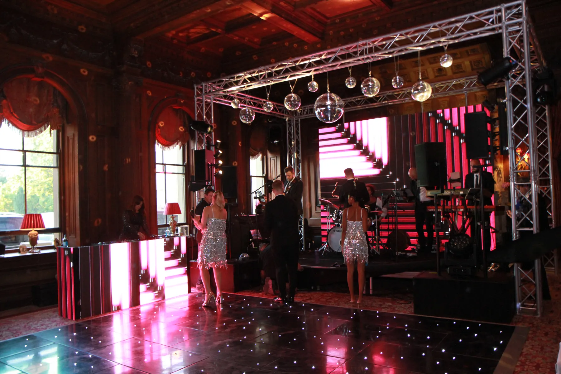 A glamorous event with people in sparkling outfits dancing on a vibrant dancefloor hire, near a stage with a live band and large LED screens in an ornately decorated room with tall windows and red curtains.