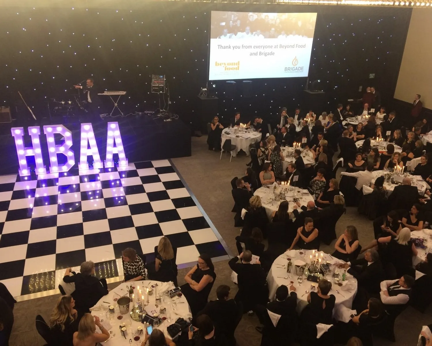 A formal event with guests seated at round tables around a black-and-white chequered dancefloor hire, large illuminated HBAA letters, and a stage; a presentation is projected on a screen at the back.