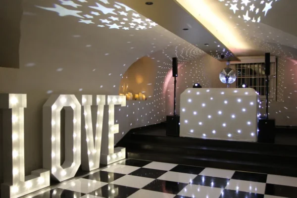 A decorative event space featuring a black-and-white chequered dancefloor hire, a DJ booth with white lights, a disco ball casting star patterns on the ceiling, and large illuminated letters spelling LOVE.
