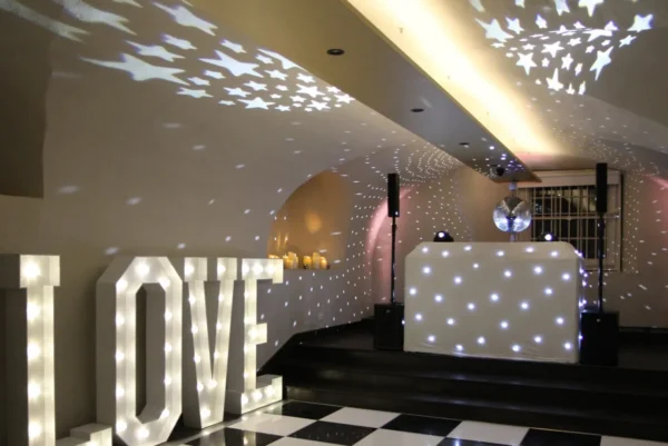 A decorative event space featuring a black-and-white chequered dancefloor hire, a DJ booth with white lights, a disco ball casting star patterns on the ceiling, and large illuminated letters spelling LOVE.