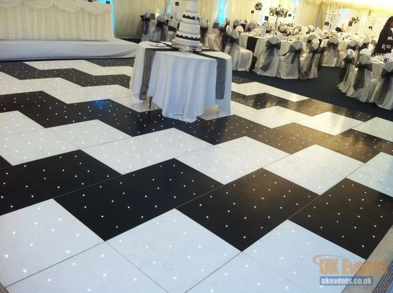 A black-and-white chequered dance floor with small embedded lights, available through dancefloor hire, set in a decorated event hall with round tables, white tablecloths, and chairs tied with dark bows. A tiered cake sits on a table in the middle.
