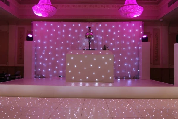 A stage with a white DJ booth and a sparkling backdrop of small lights, illuminated by pink lighting. Two pink chandeliers hang from the ceiling, while the shiny dancefloor hire option reflects the vibrant lights above.