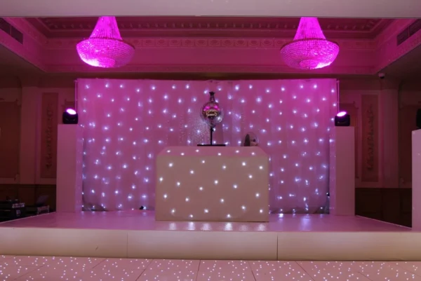 A stage with a white DJ booth and a sparkling backdrop of small lights, illuminated by pink lighting. Two pink chandeliers hang from the ceiling, while the shiny dancefloor hire option reflects the vibrant lights above.