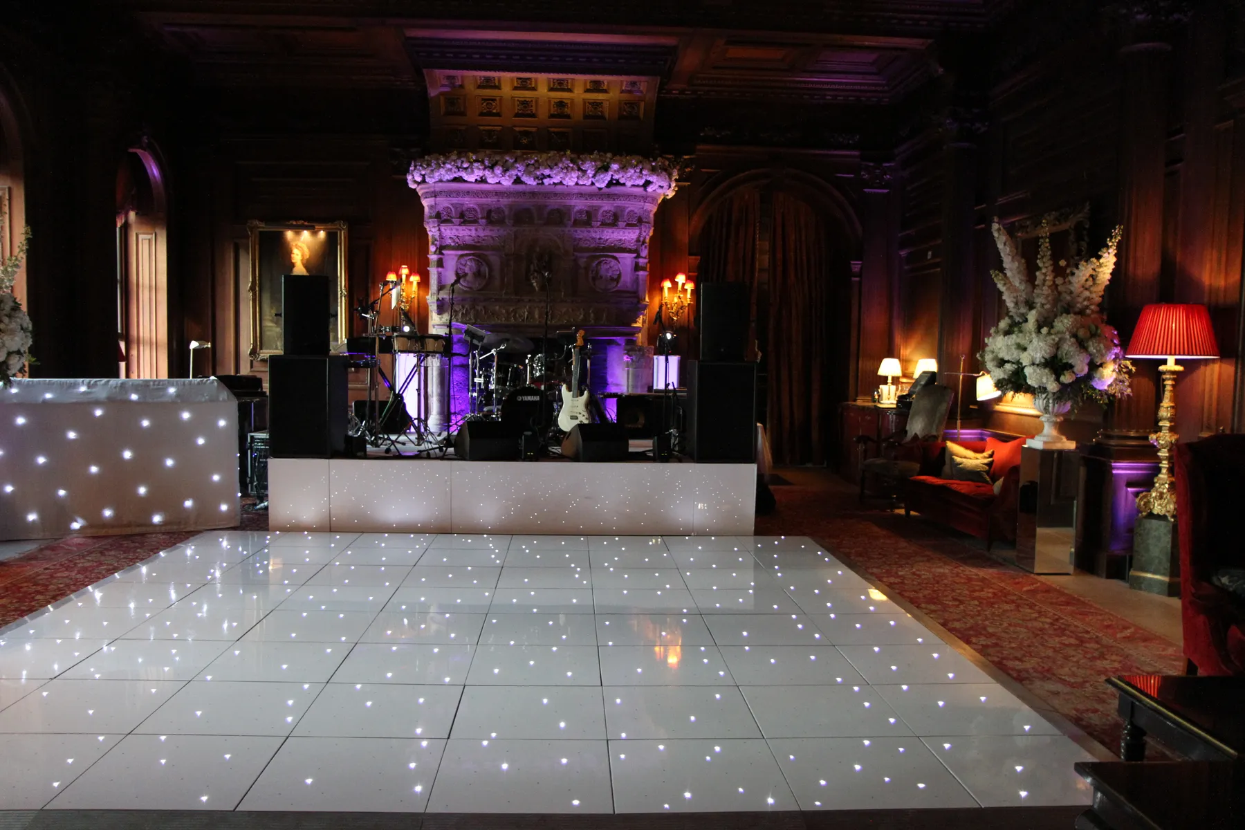 Elegant ballroom with a glossy, illuminated dance floor available for dancefloor hire, DJ booth, drum kit, ornate fireplace, dim lighting, floral arrangements, and vintage furniture in a grand, wood-panelled room.