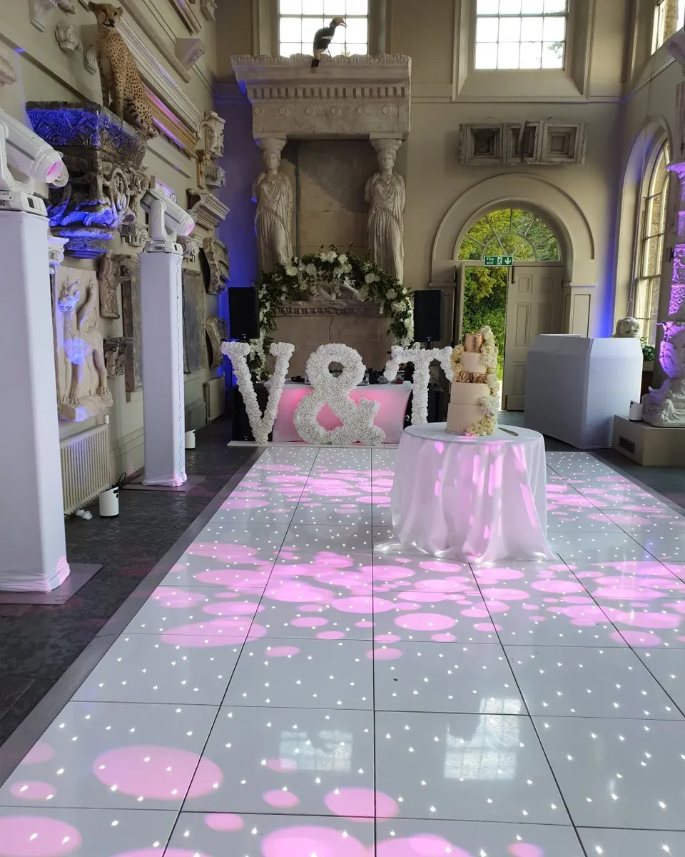 A decorated event hall with statues, a white dancefloor hire illuminated by pink and white lights, a round table with a cake, and large white floral letters V & T near a flower-draped fireplace.