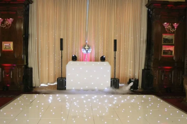 A DJ booth with a white, illuminated front panel stands in front of beige curtains, flanked by speakers and uplighters. A disco ball hangs above, and the shiny dancefloor hire is dotted with small, glowing white lights.