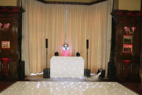A DJ booth with a white, illuminated front panel stands in front of beige curtains, flanked by speakers and uplighters. A disco ball hangs above, and the shiny dancefloor hire is dotted with small, glowing white lights.