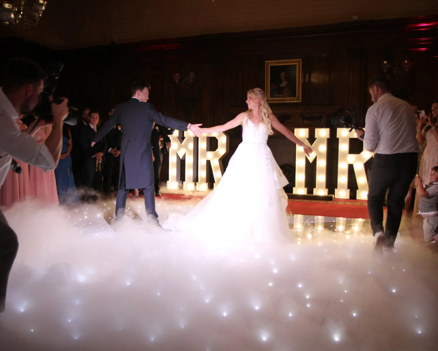 A bride and groom share their first dance on a lit-up, fog-covered floor, surrounded by guests. Large illuminated MR & MRS letters and stunning dancefloor hire create a truly romantic wedding scene.