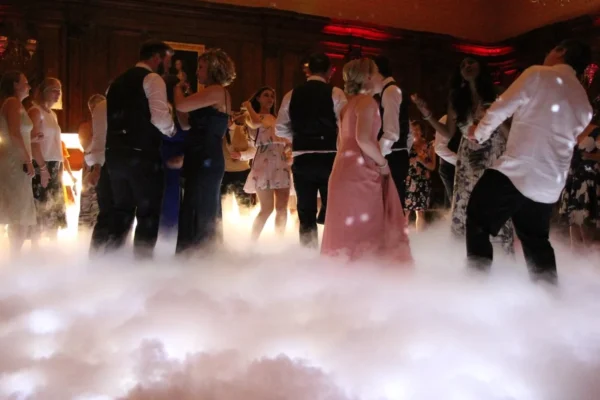 A group of people dressed in formal attire dance in a dimly lit room with wood-panelled walls, while white fog drifts across the floor—courtesy of a professional dancefloor hire—creating a dreamy, elegant atmosphere.