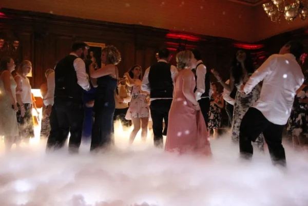 A group of people dressed in formal attire dance in a dimly lit room with wood-panelled walls, while white fog drifts across the floor—courtesy of a professional dancefloor hire—creating a dreamy, elegant atmosphere.