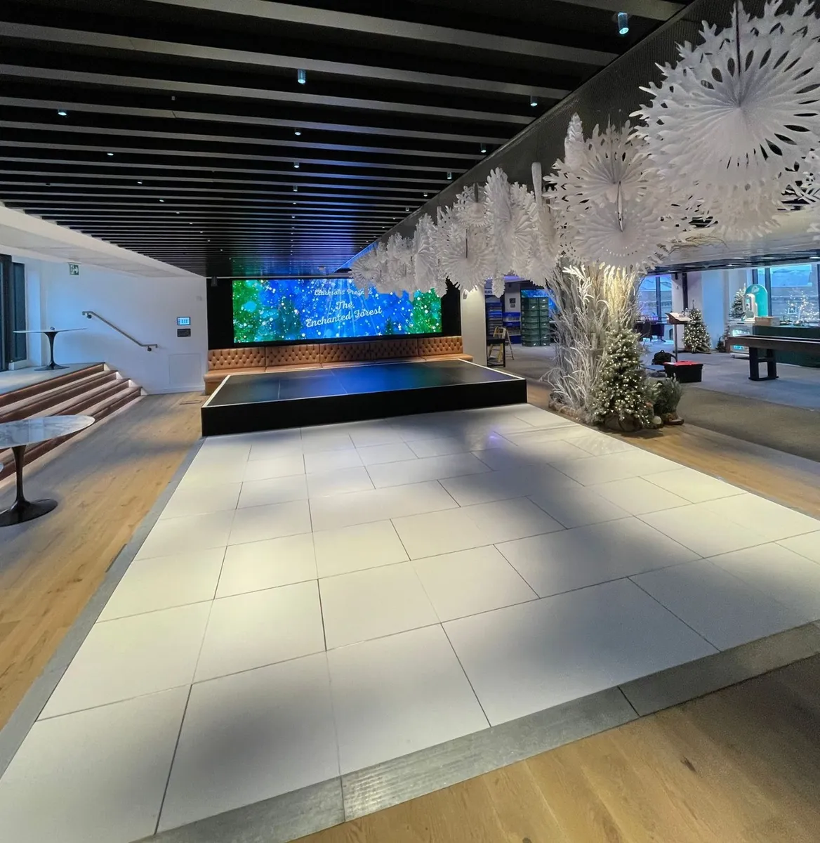 A spacious modern lobby with wooden floors perfect for dancefloor hire, a small stage, a large digital screen displaying colourful graphics, white decorative paper structures, and floor-to-ceiling windows letting in natural light.