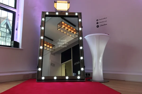 A large, upright magic mirror framed by round lights stands on a red carpet in a room with wooden floors and white walls. A tall, round white table is nearby, and a window lets in natural light.