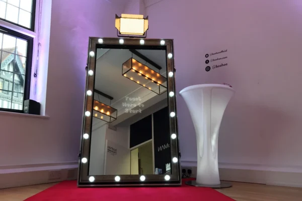 A large, upright magic mirror framed by round lights stands on a red carpet in a room with wooden floors and white walls. A tall, round white table is nearby, and a window lets in natural light.