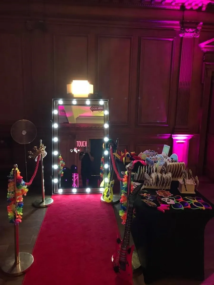 A magic mirror photo booth stands on a red carpet, surrounded by velvet ropes, colourful props like glasses and inflatable guitars, and a display table. The scene glows in pink lighting inside a dimly lit room with wood-panelled walls.