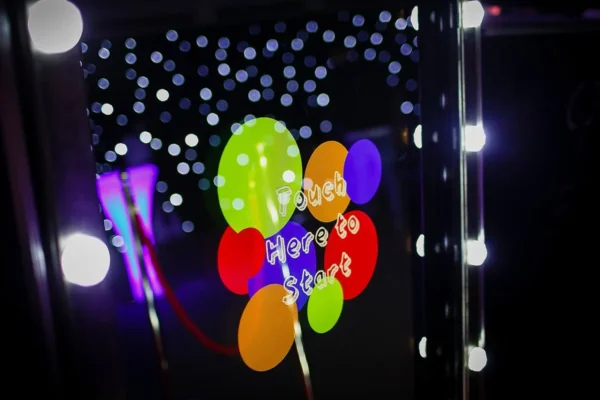 A colourful touchscreen magic mirror display with the words “Touch Here to Start,” surrounded by bright dots and illuminated by fairy lights in a dark setting, with a red velvet rope in the background.