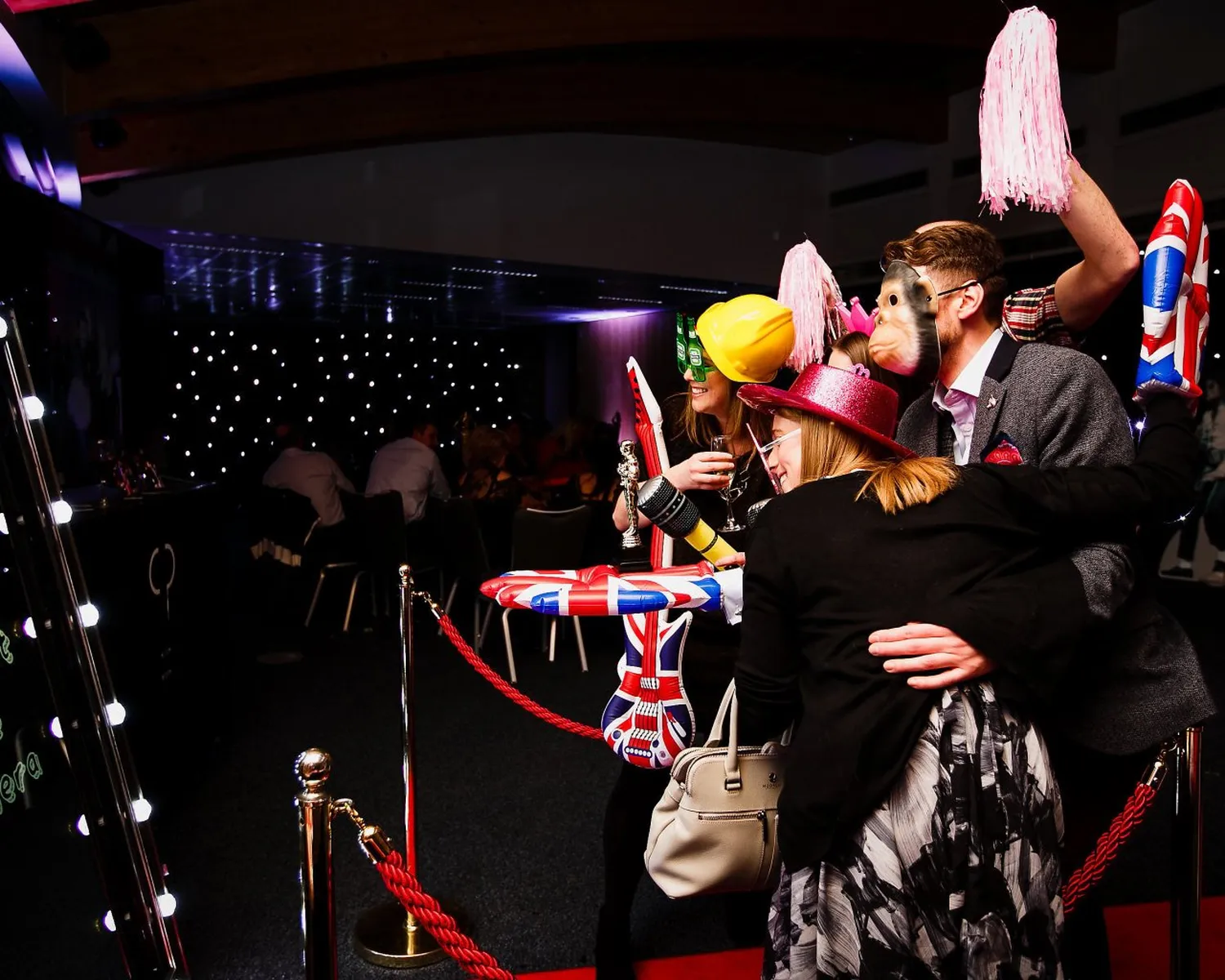A group of people in party attire pose for a photo behind a red rope, gathered round a magic mirror, wearing playful masks, hats, and holding inflatable props, with a dark, star-lit background and others seated behind them.