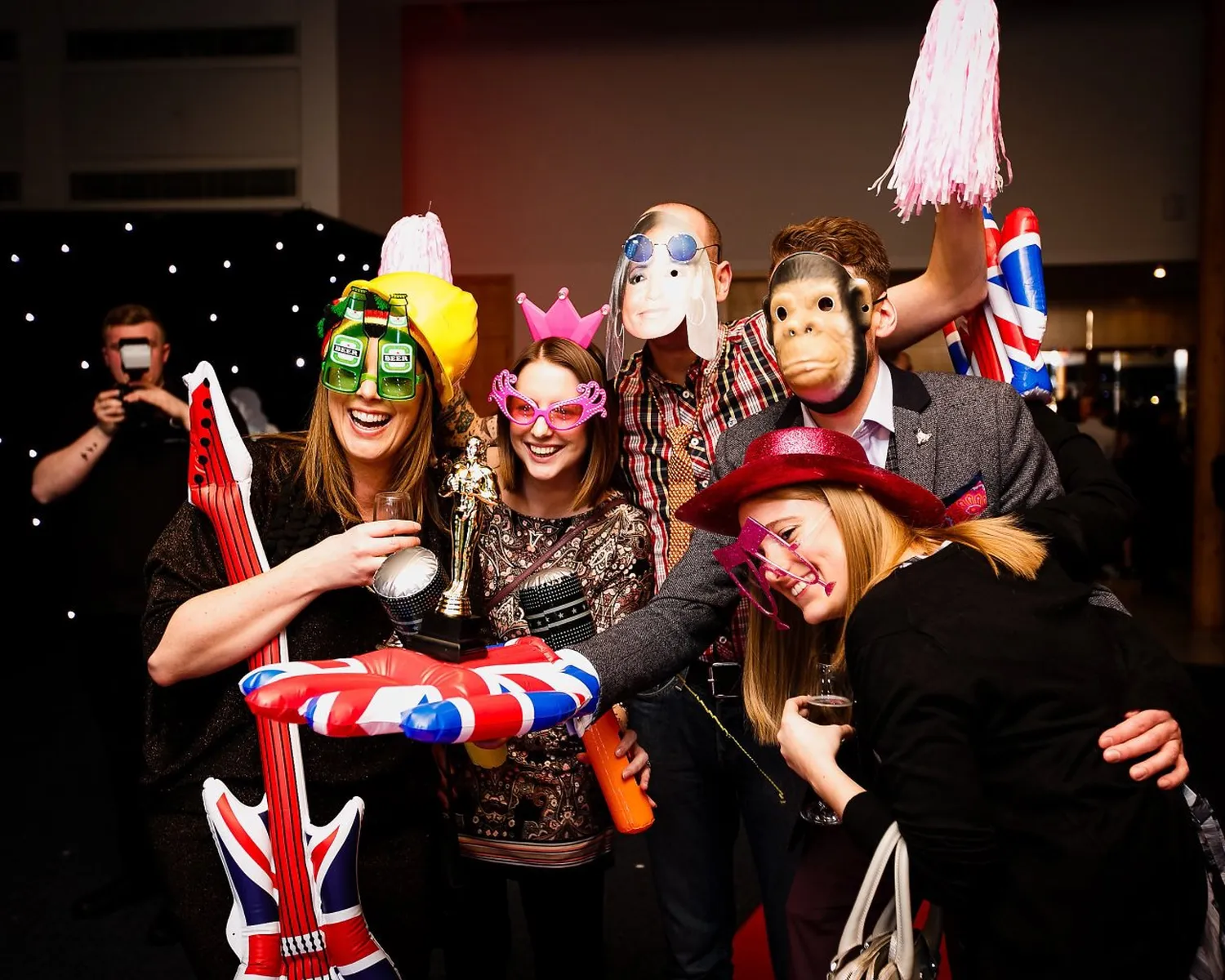 A group of people at a party pose together by the magic mirror, wearing funny masks, hats, and novelty glasses. They hold inflatable props, including a guitar and a Union Jack-themed float, whilst smiling and enjoying themselves.