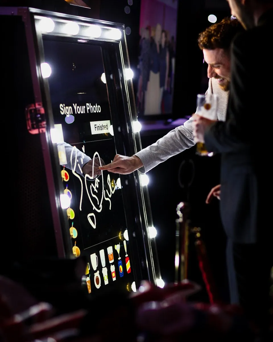 Two men interact with a brightly lit magic mirror photo booth. One man draws on the touchscreen whilst holding a drink, and the other stands beside him, partially visible. The screen says Sign Your Photo and displays digital icons.