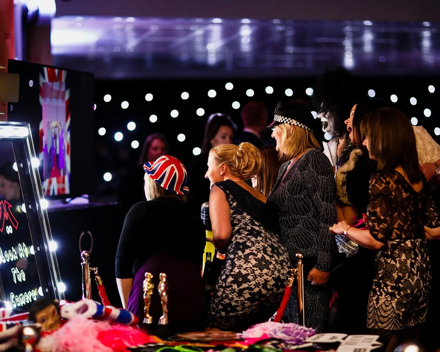 A group of women in fancy dress laugh and pose in front of a magic mirror photo booth at a lively indoor event with fairy lights in the background. Props and accessories are visible on a table nearby.