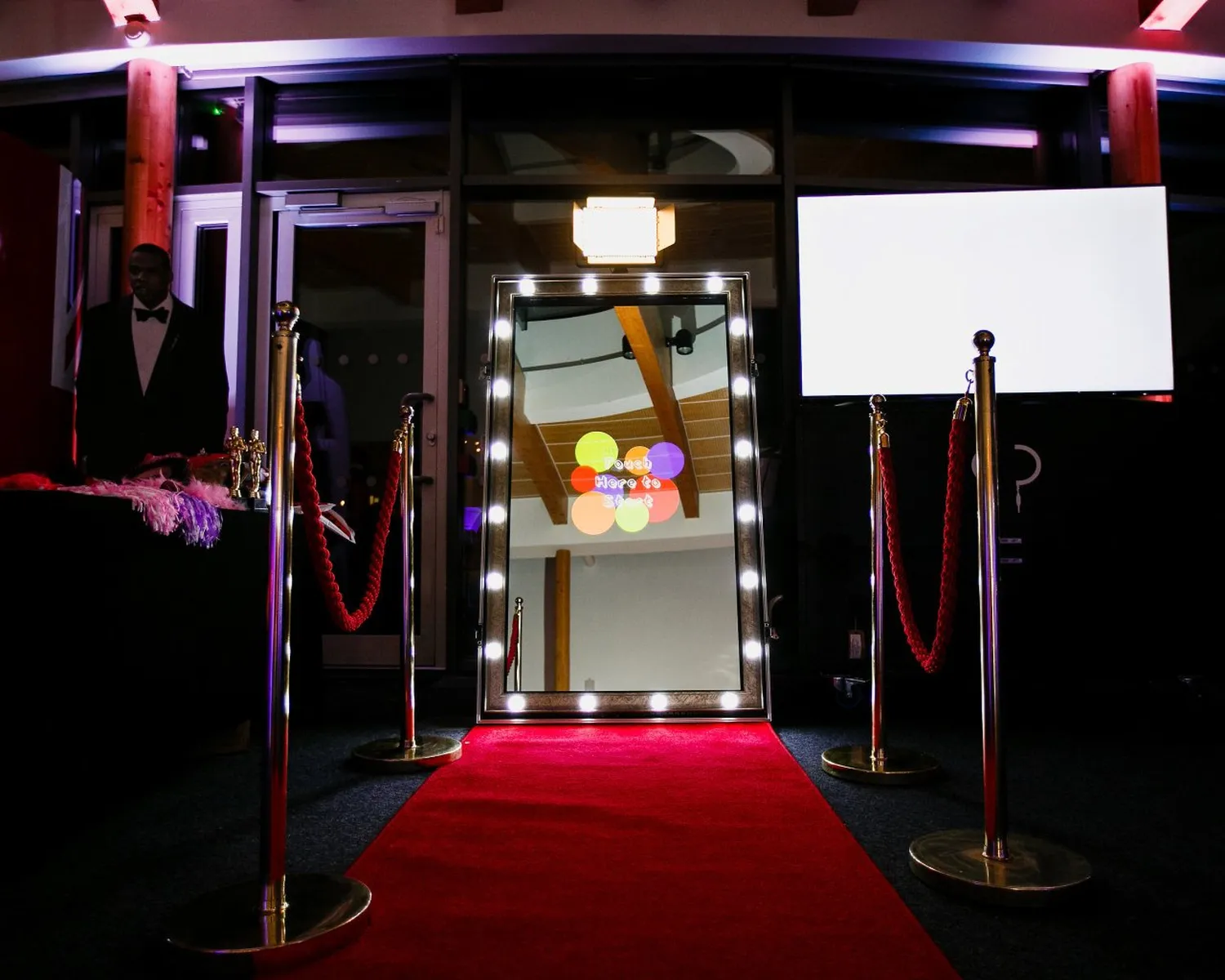 A magic mirror photo booth stands lit up at the end of a red carpet, flanked by red velvet ropes and gold posts, with a white screen and a person partially visible on the left.