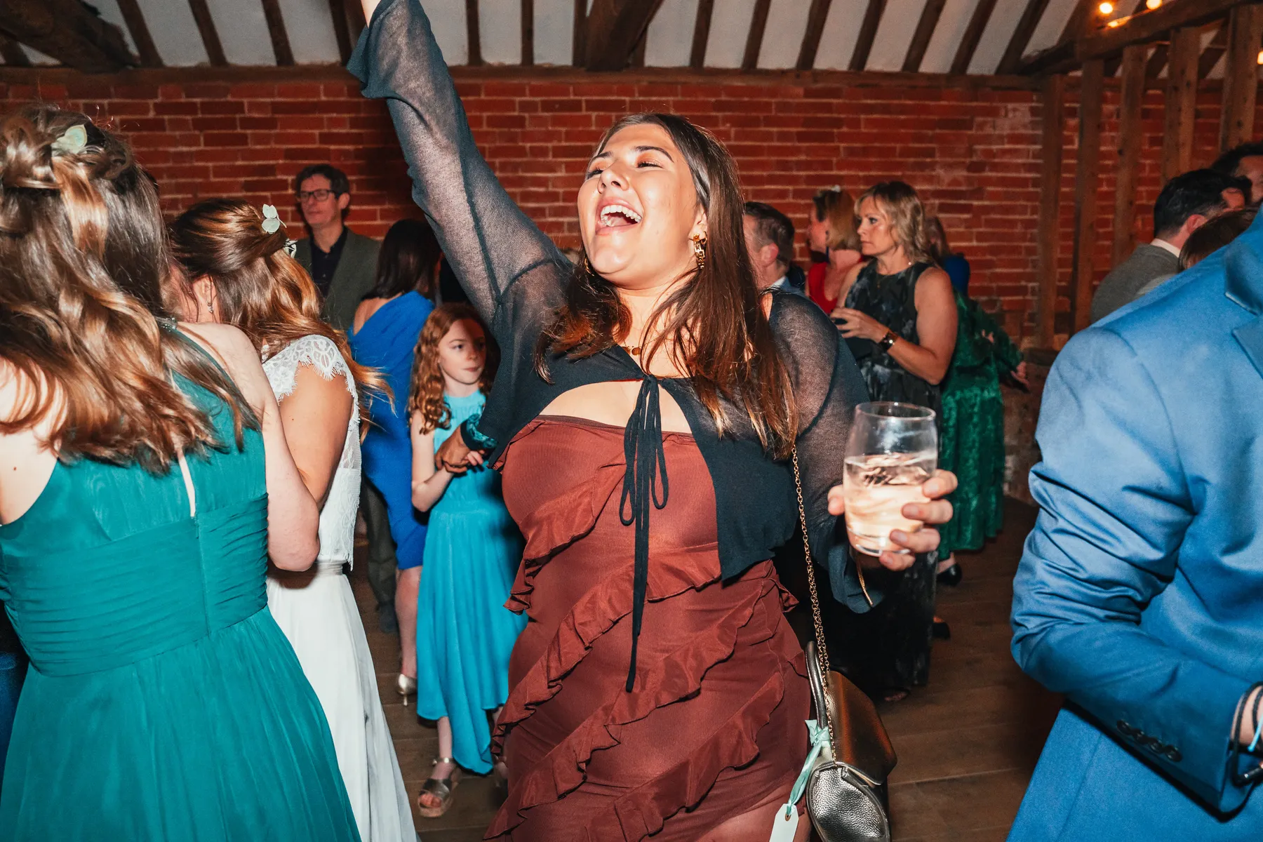 A woman in a frilled brown dress smiles and raises her arm while holding a drink, celebrating with others at a lively indoor party with a party DJ and guests dressed in formal attire.