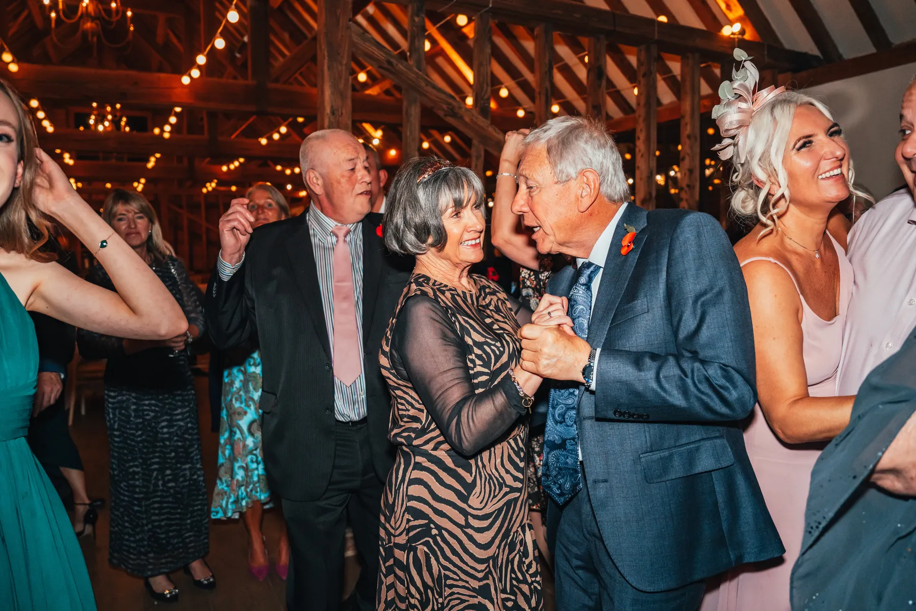 A group of well-dressed adults dance and smile together inside a warmly lit, rustic venue with wooden beams and fairy lights overhead as a party DJ keeps the energy high.