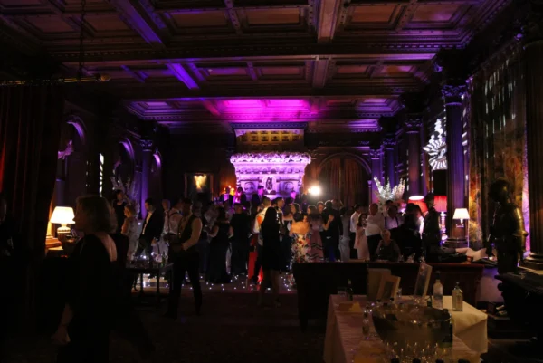 A large group of people dressed in formal attire socialise in an ornately decorated, dimly lit room with high ceilings, elegant columns, and purple mood lighting, while a party DJ sets the tone. Tables with drinks and flowers are visible in the foreground.