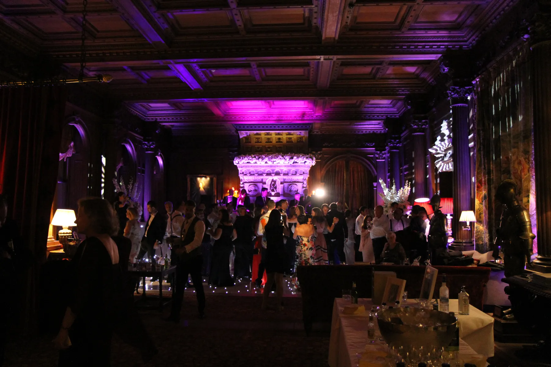 A large group of people dressed in formal attire socialise in an ornately decorated, dimly lit room with high ceilings, elegant columns, and purple mood lighting, while a party DJ sets the tone. Tables with drinks and flowers are visible in the foreground.