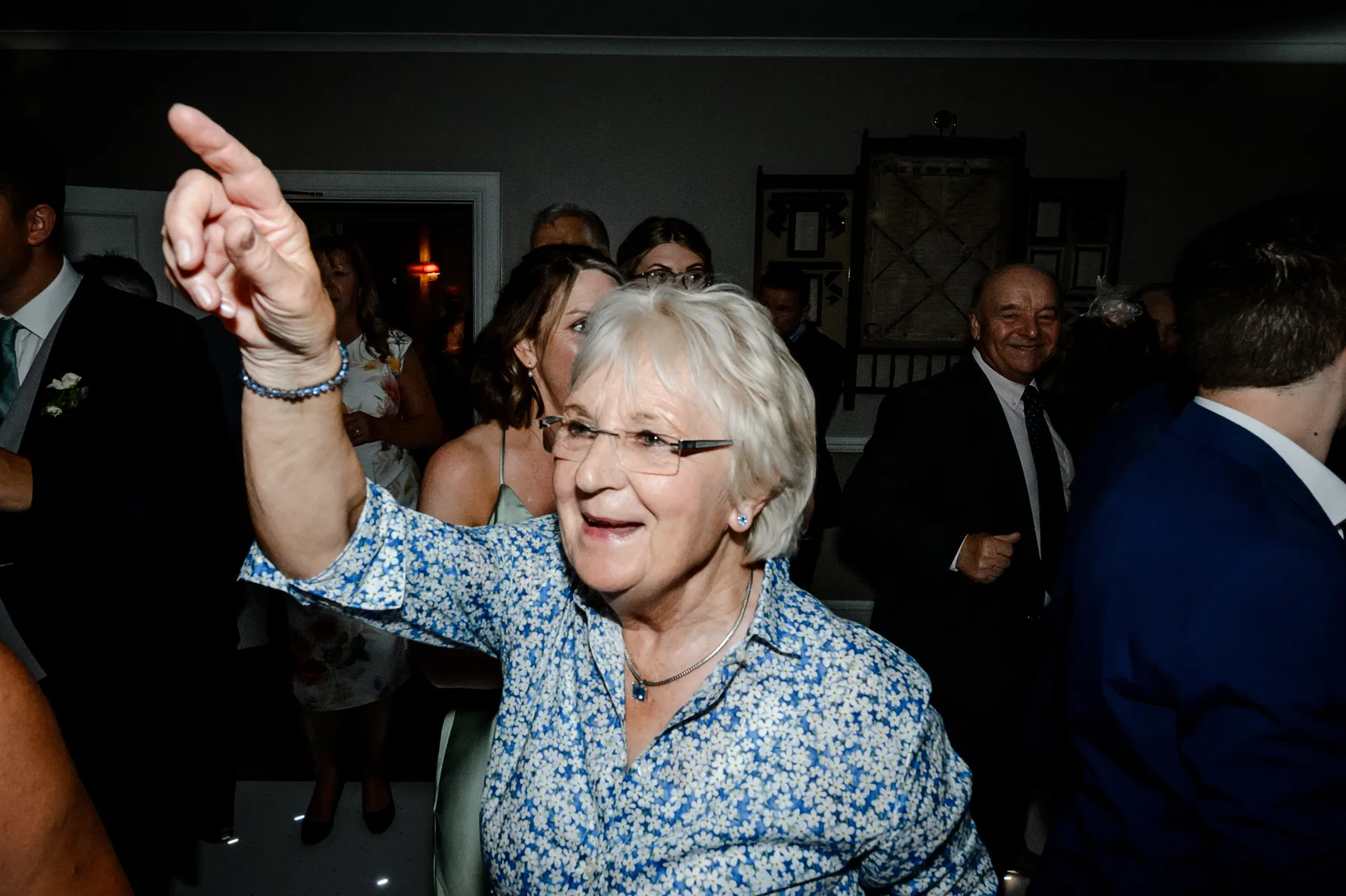An older woman with white hair and glasses, wearing a blue floral shirt, smiles and points upwards whilst dancing to the beats of a party DJ at a lively indoor gathering with other people in the background.