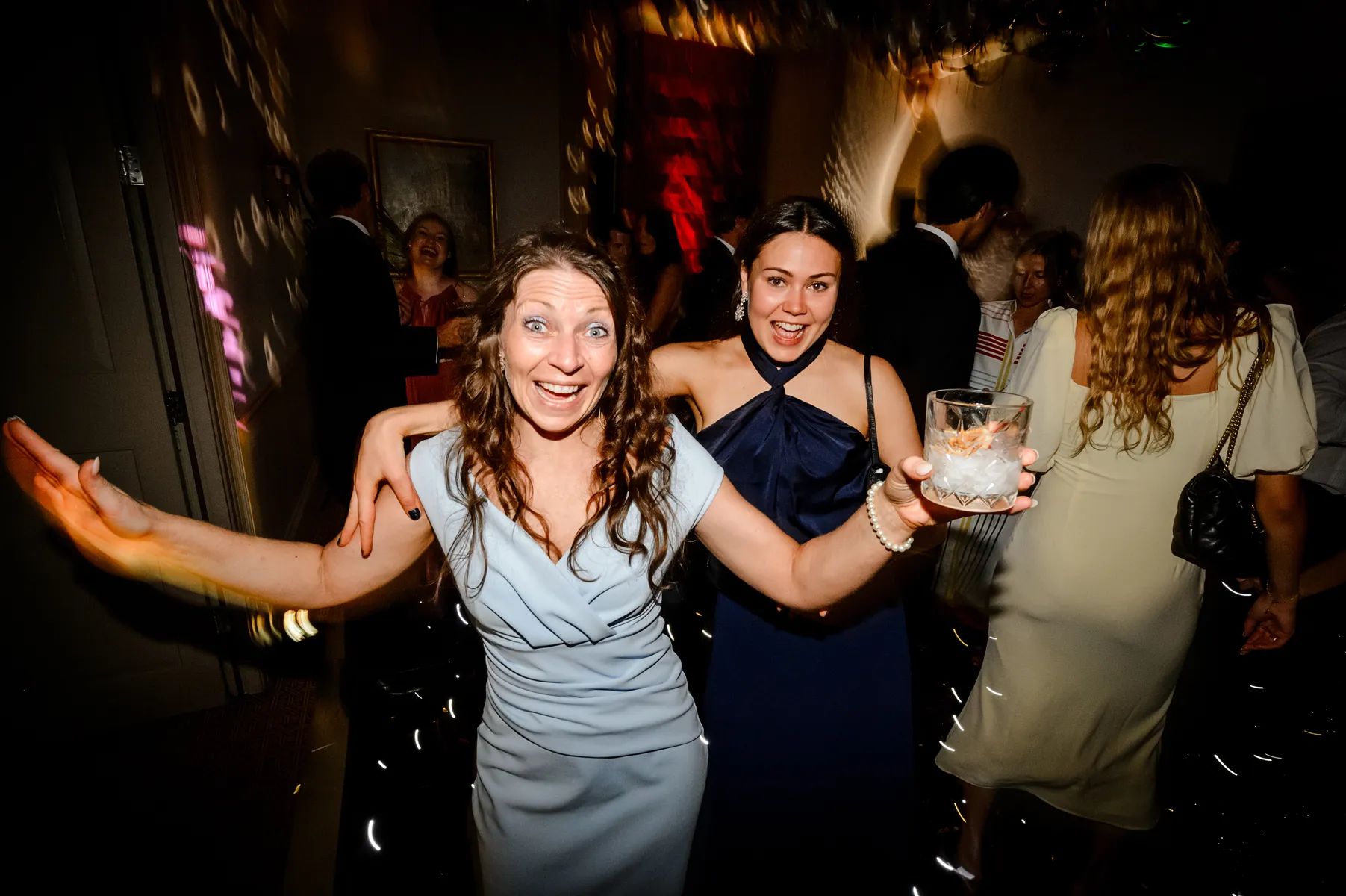 Two women smile and pose energetically on a dance floor at a party, one in a light blue dress holding a drink with ice, the other in a dark blue dress, as the party DJ spins tracks and colourful lights illuminate the dancing crowd in the background.