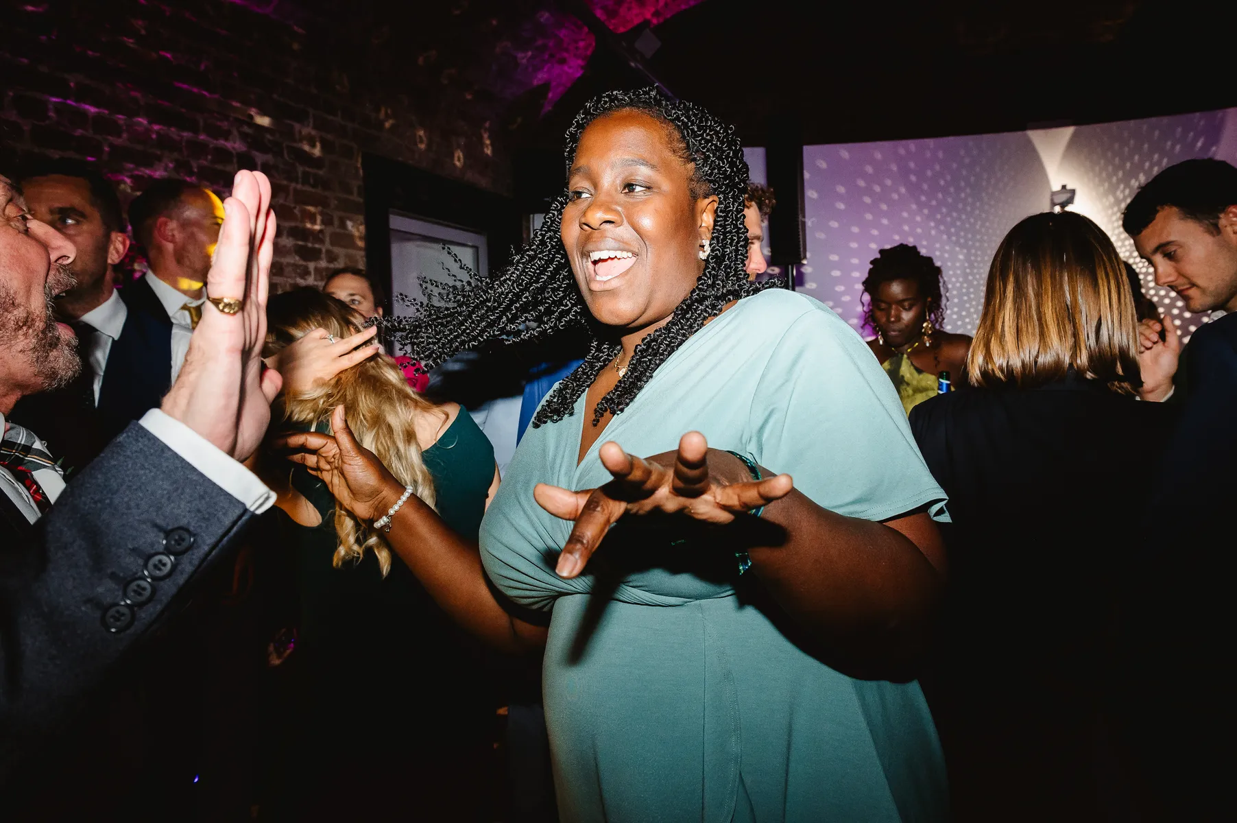 A woman in a green dress smiles and dances enthusiastically at a lively indoor party, energised by the beats of a party DJ, surrounded by others who are also celebrating and enjoying the event.