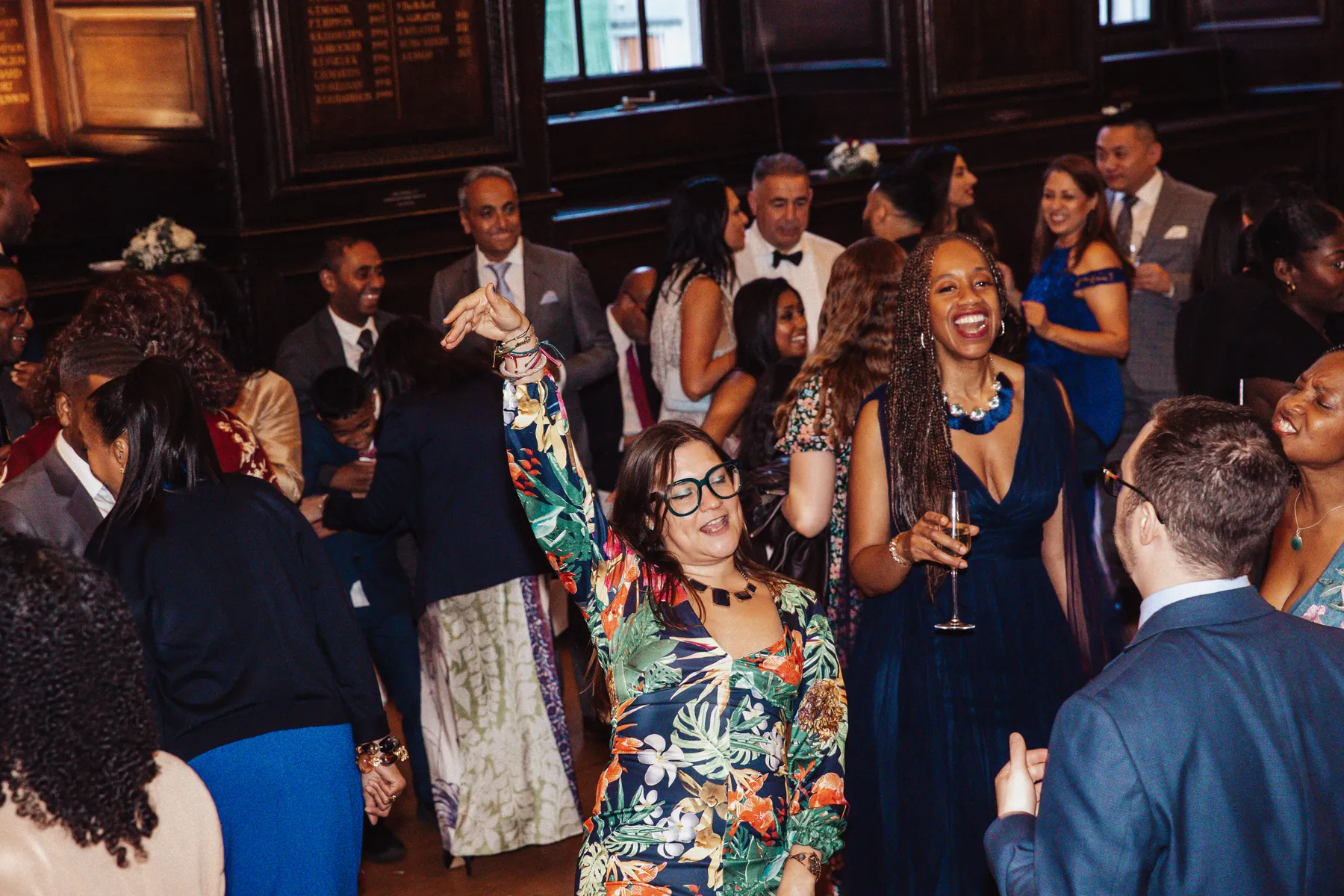 A group of well-dressed people enjoy themselves at an indoor party; one woman in a colourful dress and large glasses dances with her arm raised as a party DJ plays music, while others chat and laugh around her.