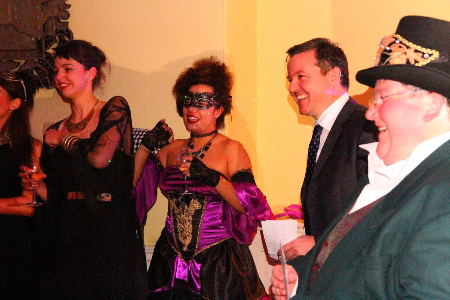A group of people in formal and masquerade attire smile and hold drinks at a festive indoor event, enjoying the lively beats from the party DJ. One wears a purple and black mask and corset; another a suit and tie, and another a top hat with gold decorations.